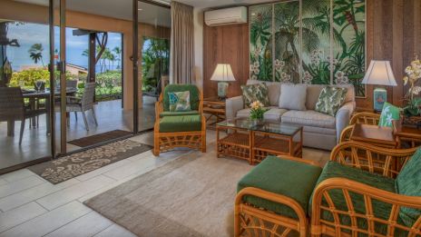 A cozy living room with wicker furniture, tropical-themed decor, and a view of an outdoor patio with palm trees visible.