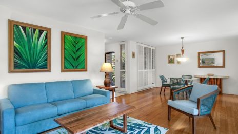 A living room with blue furniture, wooden table, ceiling fan, and tropical-themed artwork on the wall, featuring a cozy and inviting setup.