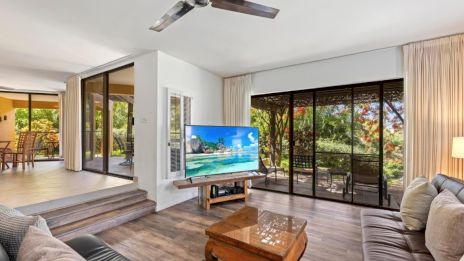 A modern living room with a TV, leather sofas, ceiling fan, wooden table, and large glass doors opening to a lush outdoor view.