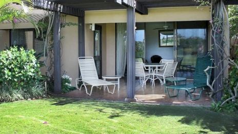 A patio with outdoor furniture, including lounge chairs and a table with chairs, under a pergola. Lush green lawn in the foreground.