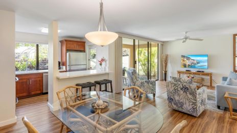The image shows a modern, open-plan kitchen and living area with wooden floors, glass table, floral chairs, and a TV.