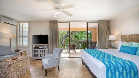 A bright room with a bed, TV, and sitting area, featuring a ceiling fan, large windows, and access to a balcony with outdoor seating.