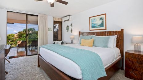 A cozy bedroom with a large bed, blue bedding, a ceiling fan, and balcony view with outdoor seating. Decor includes a landscape painting.