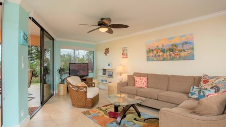 A cozy living room with a beige sofa, colorful pillows, a glass coffee table, and a TV; sliding glass doors open to a patio.