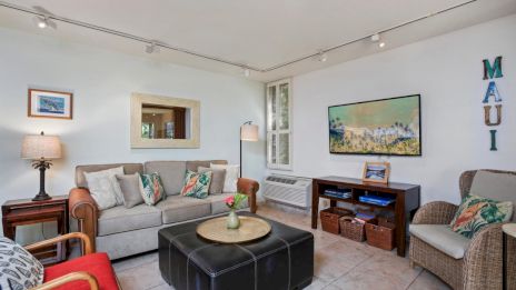 A cozy living room features a sofa, armchair, wall art, and decor with "MAUI" letters. There's a central ottoman and a side table lamp.