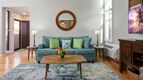 A cozy living room with a teal sofa, green pillows, wooden coffee table, wall mirror, two lamps, floral rug, and tropical-themed TV screen.