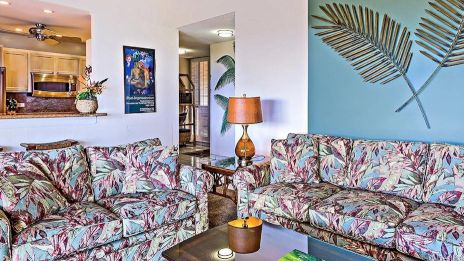 A living room features floral-patterned sofas, a coffee table, a lamp, and wall decor of metal leaves with a view into a kitchen.