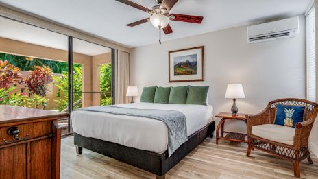 A cozy bedroom with a bed, armchair, lamps, artwork, and a ceiling fan. Large sliding doors open to a lush outdoor garden.