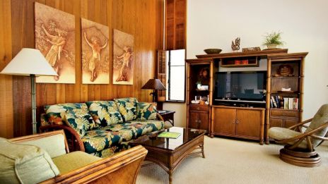 A cozy living room with wooden furniture, a floral sofa, TV unit, artistic wall panels, and warm lighting.