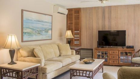 A cozy living room with a beige sofa, two lamps, a television, and a wall-mounted painting, complemented by wooden and wicker furniture.