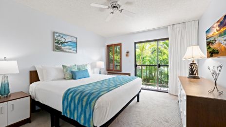A bright bedroom with a king-sized bed, blue accents, lamps, and artwork, opens to a balcony with a view of greenery.