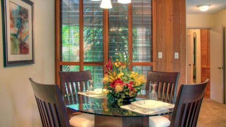 A dining area with a glass table, wooden chairs, and a floral centerpiece, next to a window with blinds and artwork on the wall.