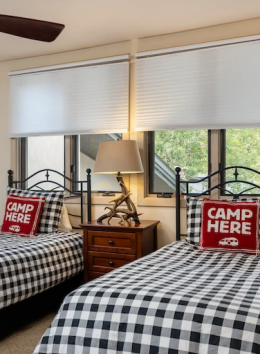 A bedroom with two twin beds, checkered bedding, pillows saying "Camp Here," a nightstand, lamp, ceiling fan, and mirrored closet.
