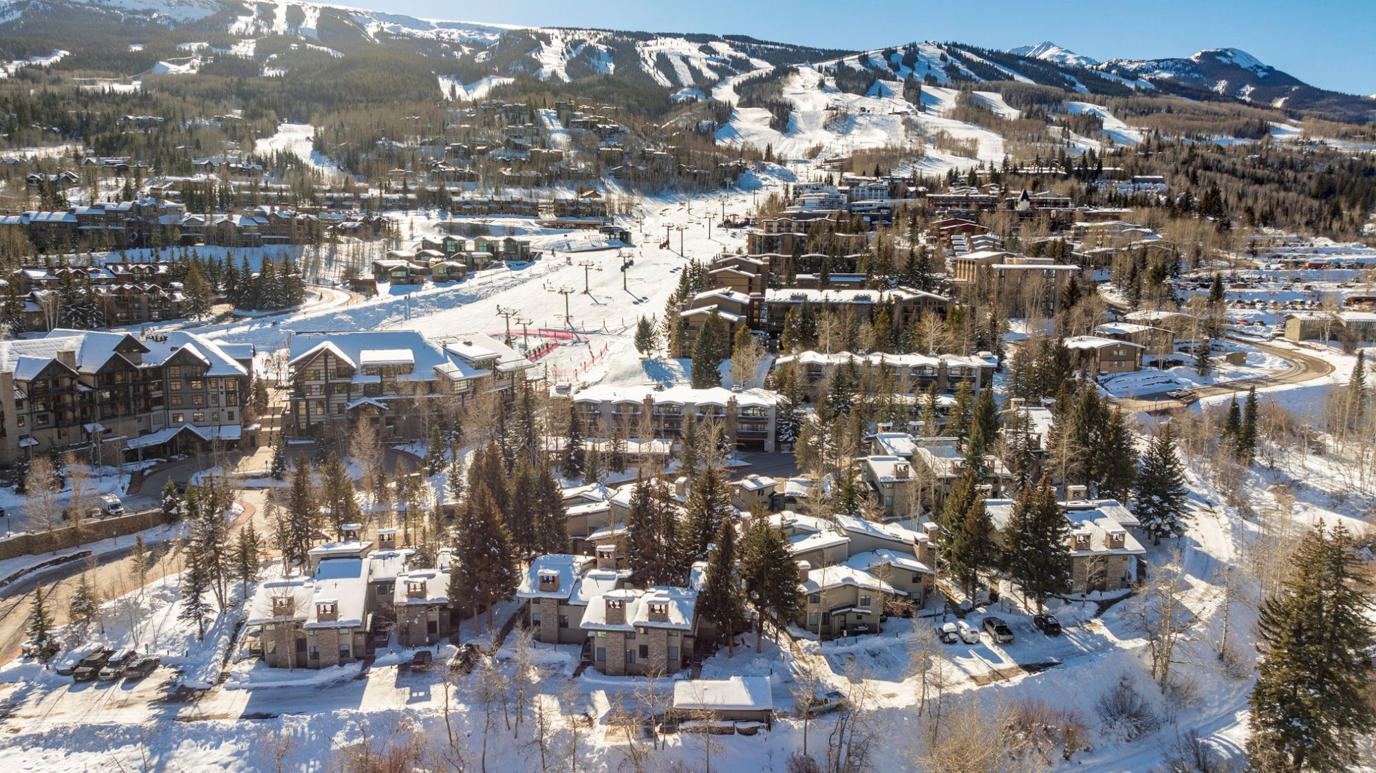 The image shows a snowy mountain resort with ski slopes, lodgings, trees, and ski lifts under a clear blue sky. It looks picturesque.