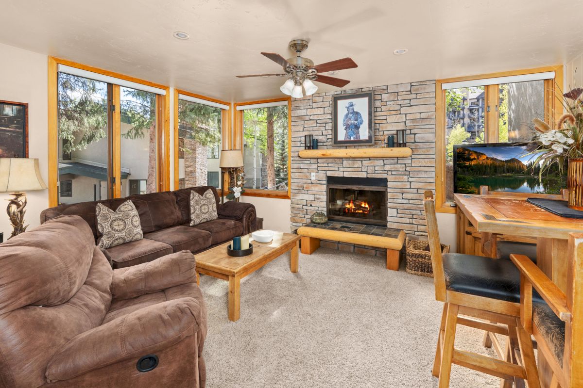 The image shows a cozy living room with a stone fireplace, brown couches, large windows, and a wooden table by the view.