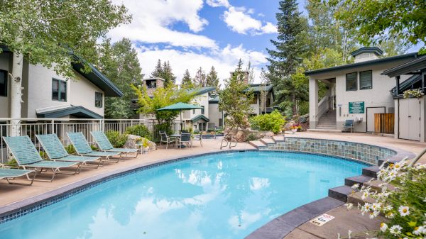 The image features a tranquil poolside scene with loungers, lush greenery, and charming buildings under a blue sky.