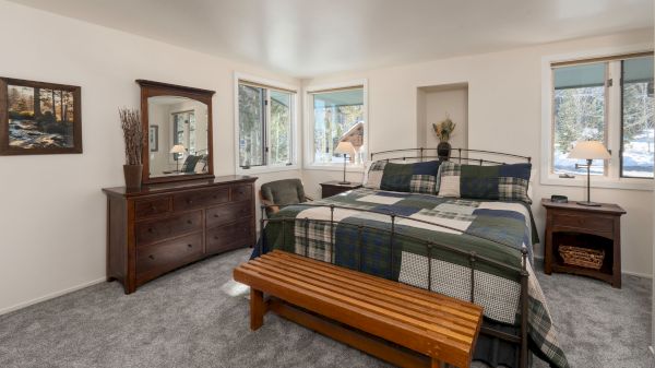 A cozy bedroom with a checkered bedspread, wooden furniture, and carpeted floor. Large windows allow natural light to fill the room.