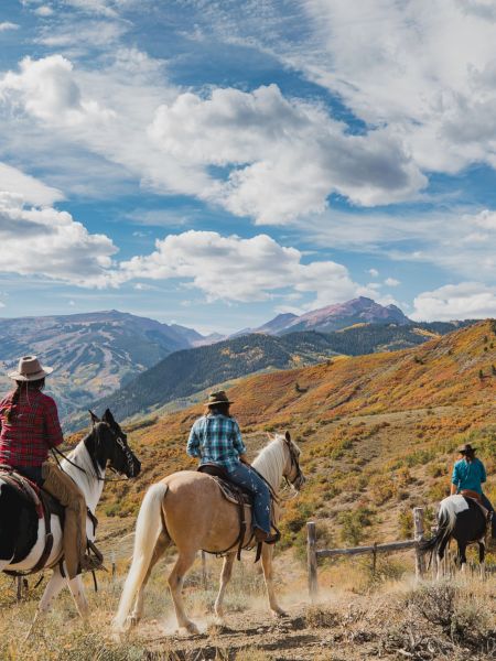Three riders on horseback navigate a scenic mountain trail, surrounded by vibrant fall foliage and expansive blue skies.