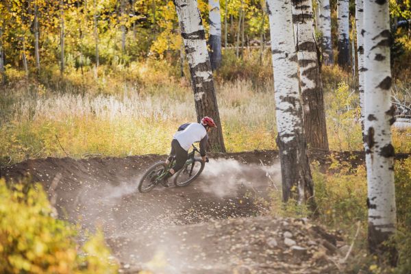 A mountain biker leans into a turn on a dirt trail surrounded by vibrant autumn foliage and aspen trees. Dust rises behind.