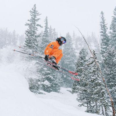 A skier in an orange outfit is jumping off a snowy slope, surrounded by tall trees in a wintery landscape. Snow is falling.