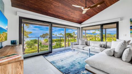 A modern living room with large windows, a sectional sofa, a wooden ceiling, and a view of greenery and water outside.