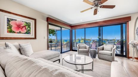 A modern living room features a sectional sofa, two chairs, a coffee table, large windows with a view, a ceiling fan, and floral artwork.