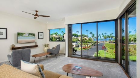 A modern living room with large windows overlooks a lush garden, featuring a TV, stylish furniture, and a ceiling fan.