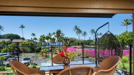 A patio with wicker chairs and a table overlooks a lush landscape with palm trees, flowers, and a clear blue sky in the background.