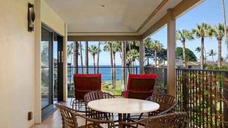 A balcony features a round table with wicker chairs, red-cushioned seats, and a view of palm trees and the ocean in the background.
