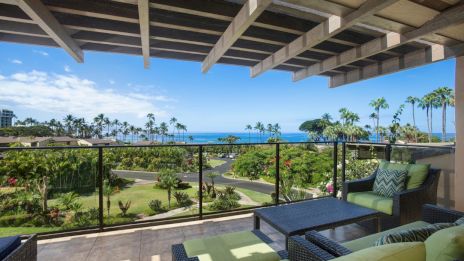 A balcony with comfy seating overlooks lush greenery, palm trees, and a distant ocean view under a partly cloudy sky.