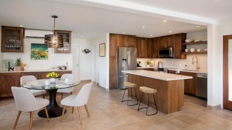The image shows a modern kitchen with wooden cabinets, a central island, stainless steel appliances, and a dining area with a round table.