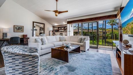 A modern living room with a white sectional sofa, patterned chair, wooden table, large rug, ceiling fan, and glass doors leading outside.