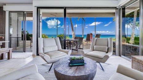 Modern living room with white chairs, round table, and large glass doors opening to a patio with ocean view and blue sky.