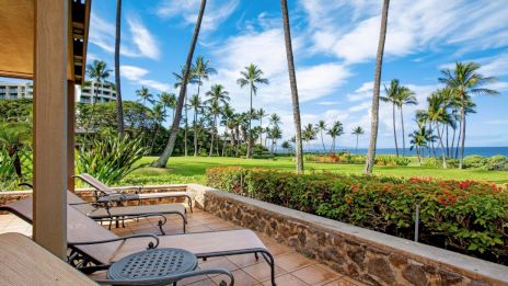 A patio view with lounge chairs overlooks a lush garden, palm trees, and an ocean under a blue sky with wispy clouds.