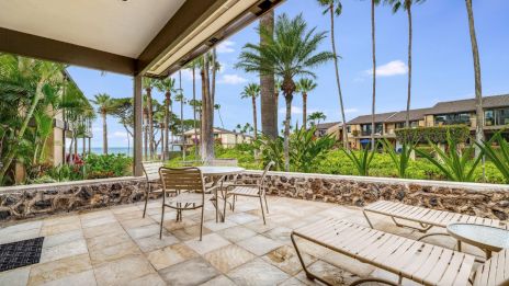 A patio with chairs and loungers overlooks a tropical garden with palm trees, leading to a view of the ocean under a clear blue sky.