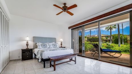 A bright bedroom with a bed, ceiling fan, and sliding glass doors opening to a patio with loungers and a garden view.