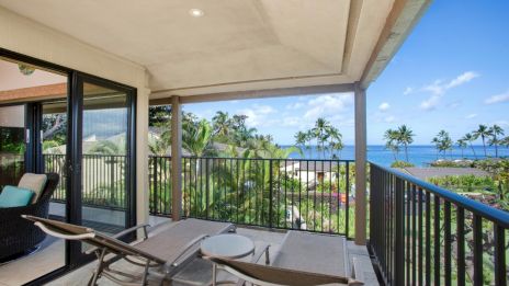 A cozy balcony with lounge chairs, a table, ocean view, and palm trees, surrounded by lush greenery under a blue sky.