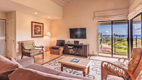 A cozy living room with a sofa, chairs, TV, and a view of the ocean through sliding glass doors, decorated with a light rug and artwork.