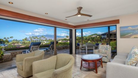 A modern living room with open views of a scenic landscape with palm trees and mountains, featuring cozy seating and a ceiling fan.