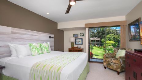 A bedroom features a large bed, green-accented decor, an armchair, and a view of greenery through sliding glass doors.
