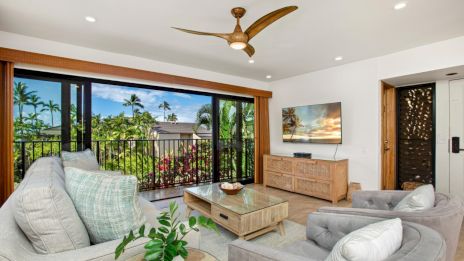 A modern living room with gray seating, wooden furniture, tropical views through large windows, and a ceiling fan on white walls.