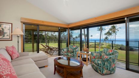 A cozy living room with a sectional sofa, patterned chairs, and a glass coffee table overlooks a scenic view of palm trees and the ocean.