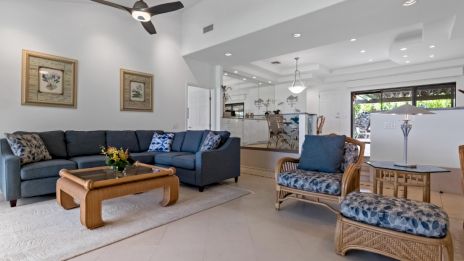 A modern living room with a blue sectional sofa, wicker chairs, a coffee table, ceiling fan, and open kitchen area in view.