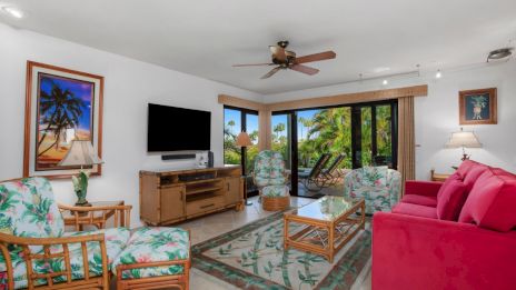 A cozy living room with a pink sofa, tropical-patterned chairs, ceiling fan, TV, and large windows showcasing a lush outdoor view.