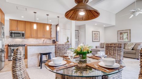 A modern open-plan living area features wicker chairs, a glass dining table, and a kitchen with wooden cabinets and pendant lights.