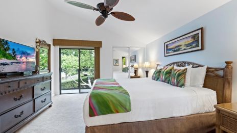 A spacious bedroom with a large bed, vibrant pillows, dresser, TV, ceiling fan, and a sliding glass door leading outside.