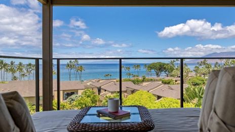 A balcony with comfy seating, a picturesque ocean view, palm trees, and a table holding a mug and a book, under a partly cloudy sky.