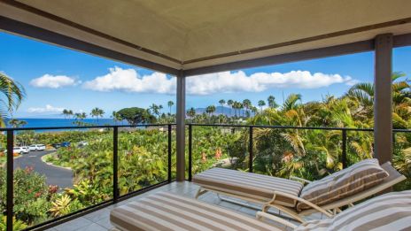 A covered balcony with two striped lounge chairs overlooks a lush landscape and ocean view, featuring palm trees and a clear blue sky.