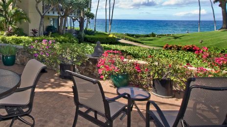 A scenic patio with chairs and tables overlooks the ocean, surrounded by vibrant flowers and a lush green lawn under a clear blue sky.