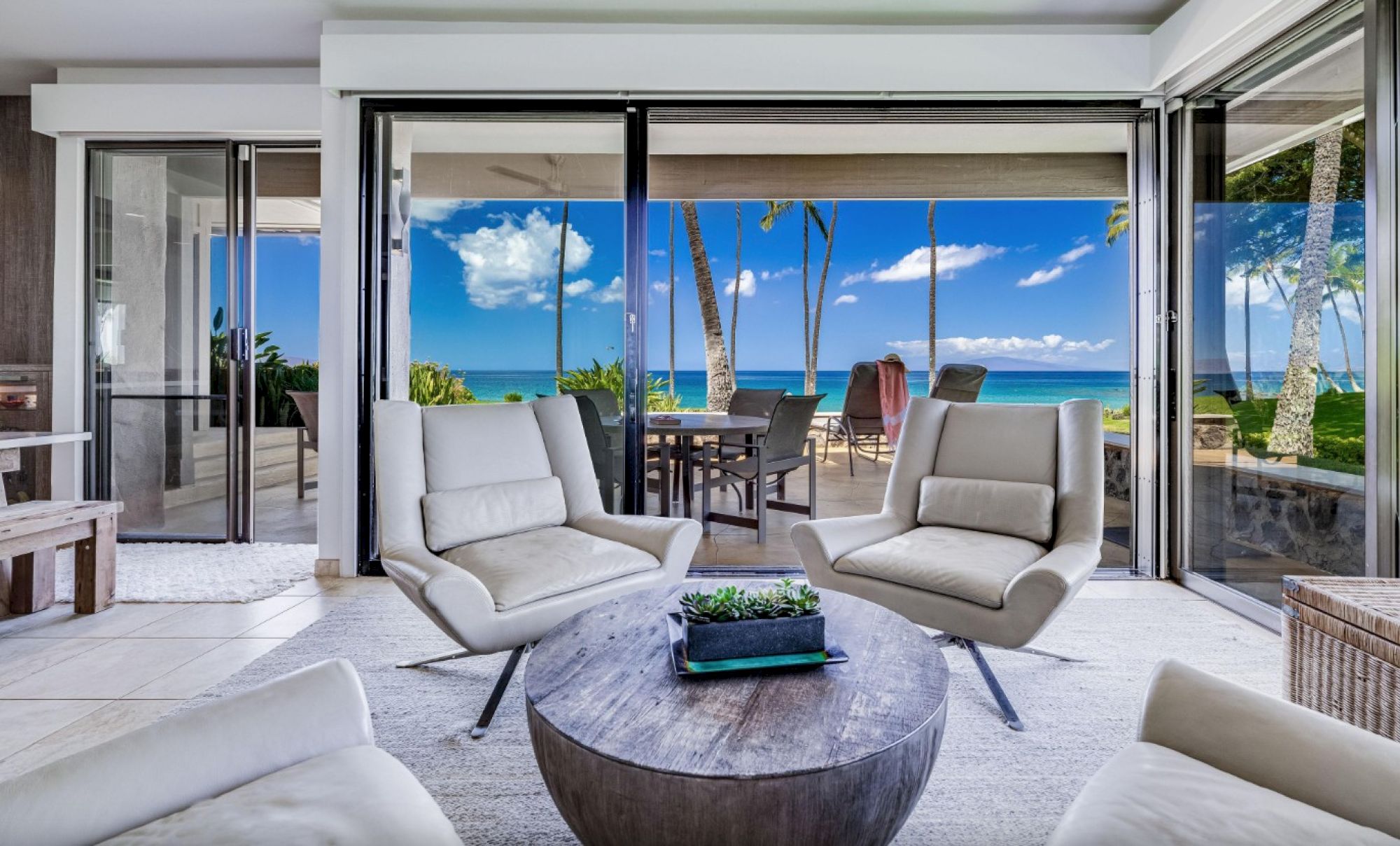 Bright, modern living room with white chairs, circular table, and glass doors leading to a patio overlooking a beach and palm trees.